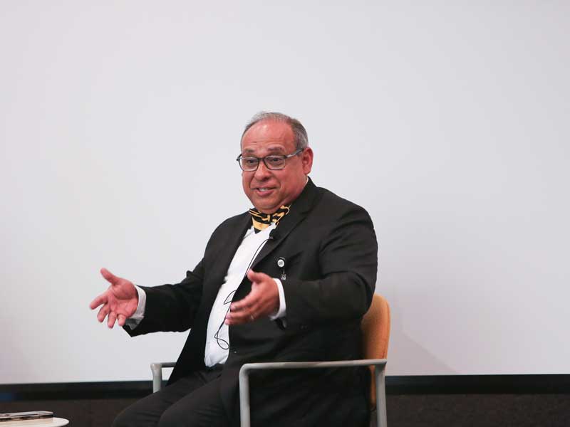 A smiling Provost Saavedra, in a suit with a black and gold bow tie, gestures toward the audience to emphasize a point while onstage at the School of Pharmacy's 4th Annual Convocation.