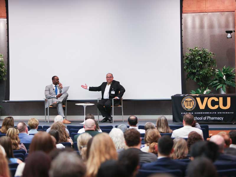 Provost Saavedra, in a suit with a black and gold bow tie, gestures toward Dean Ogbonna for emphasis while onstage next to a VCU School of Pharmacy banner at the Library of Virginia.