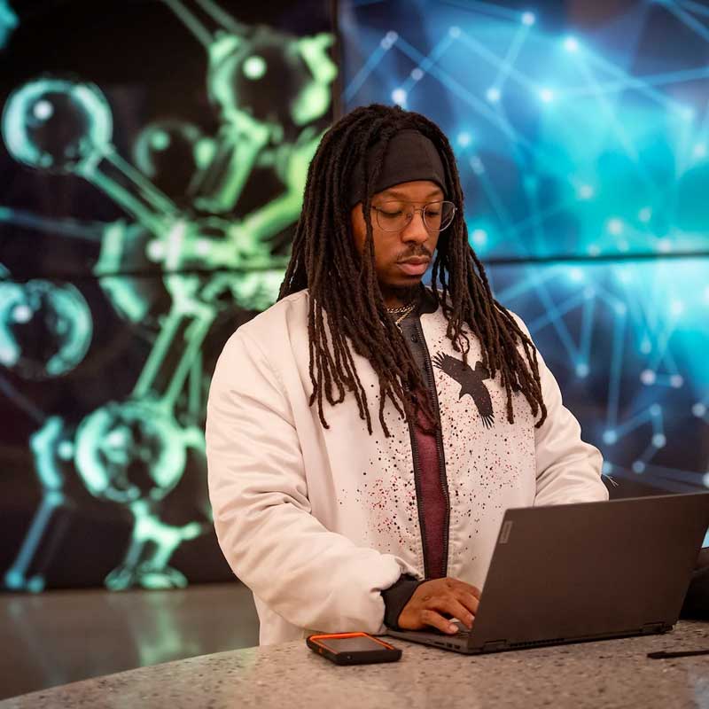 a student working on a laptop in front of a brightly colored wall featuring large displays of science images such as d.n.a. strands and molecules