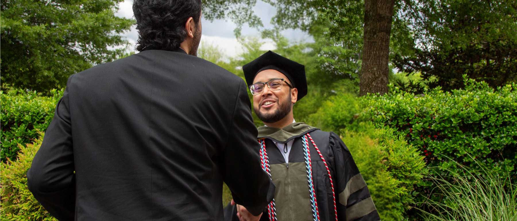 A graduate in cap and gown smiles while shaking hands with another person at an outdoor ceremony