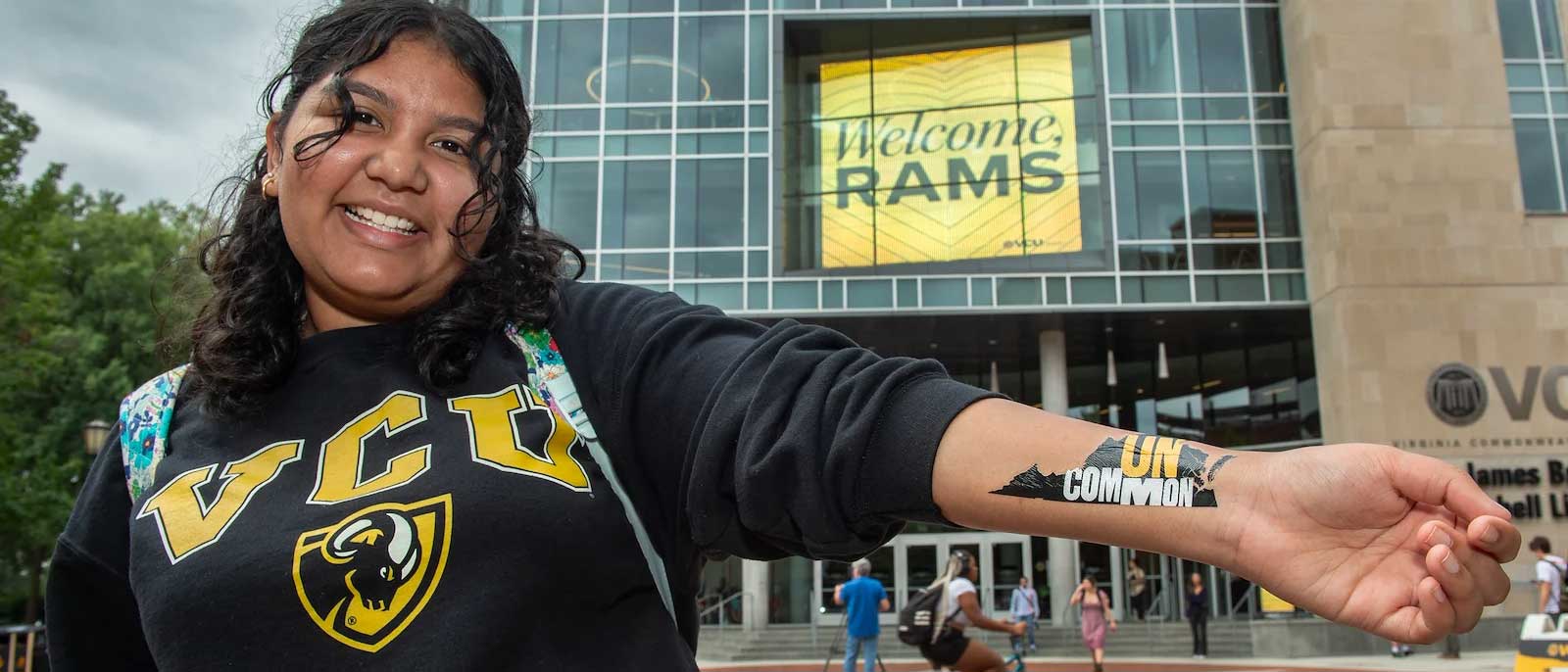 a smiling student standing outside the v.c.u. library wearing a v.c.u. sweatshirt shows off an arm tattoo in the shape of the state of virginia that says 'uncommon'