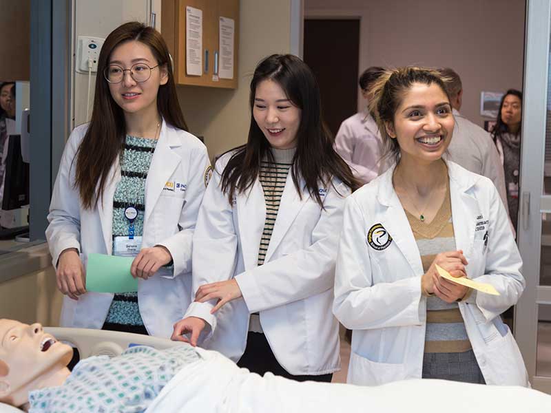 Three students stand next to a hospital bed with a human cadaver.
